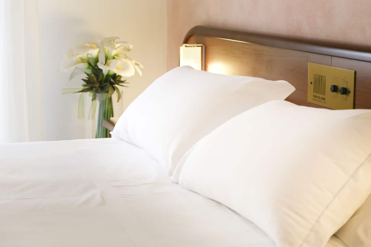 A hotel room with a neatly made bed, a nightstand with a vase of flowers, and a control panel on the wall.