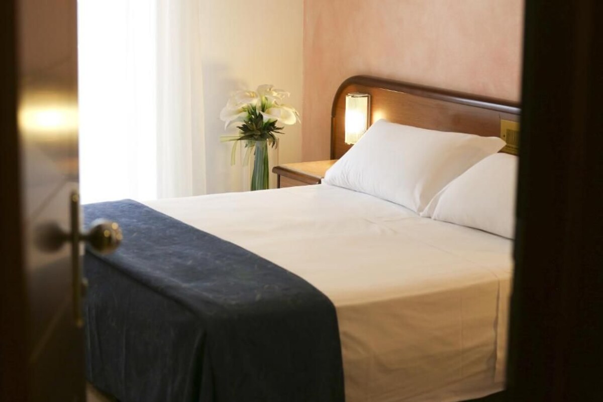 Una habitación de hotel con una cama bien hecha, una mesita de noche con lámpara y un florero con flores.