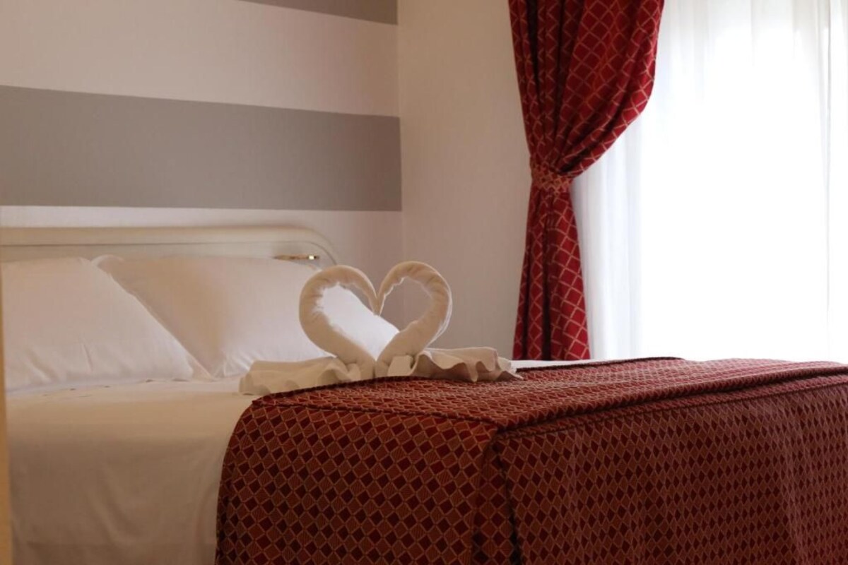 A hotel room with a neatly made bed, a heart-shaped decoration on the bed, and a window with red curtains.
