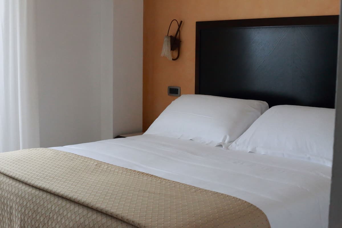 A neatly made bed with white linens and a beige blanket, a dark headboard, and a wall-mounted light fixture.