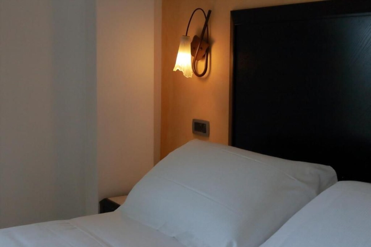 Una habitación de hotel con una cama, una lámpara de pared y un cabecero oscuro.