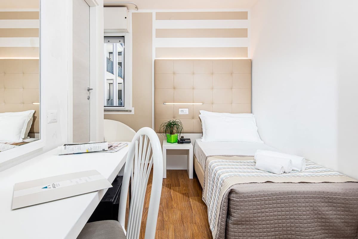 Una habitación de hotel pequeña con cama, mesita de noche, silla y un escritorio con otra silla.
