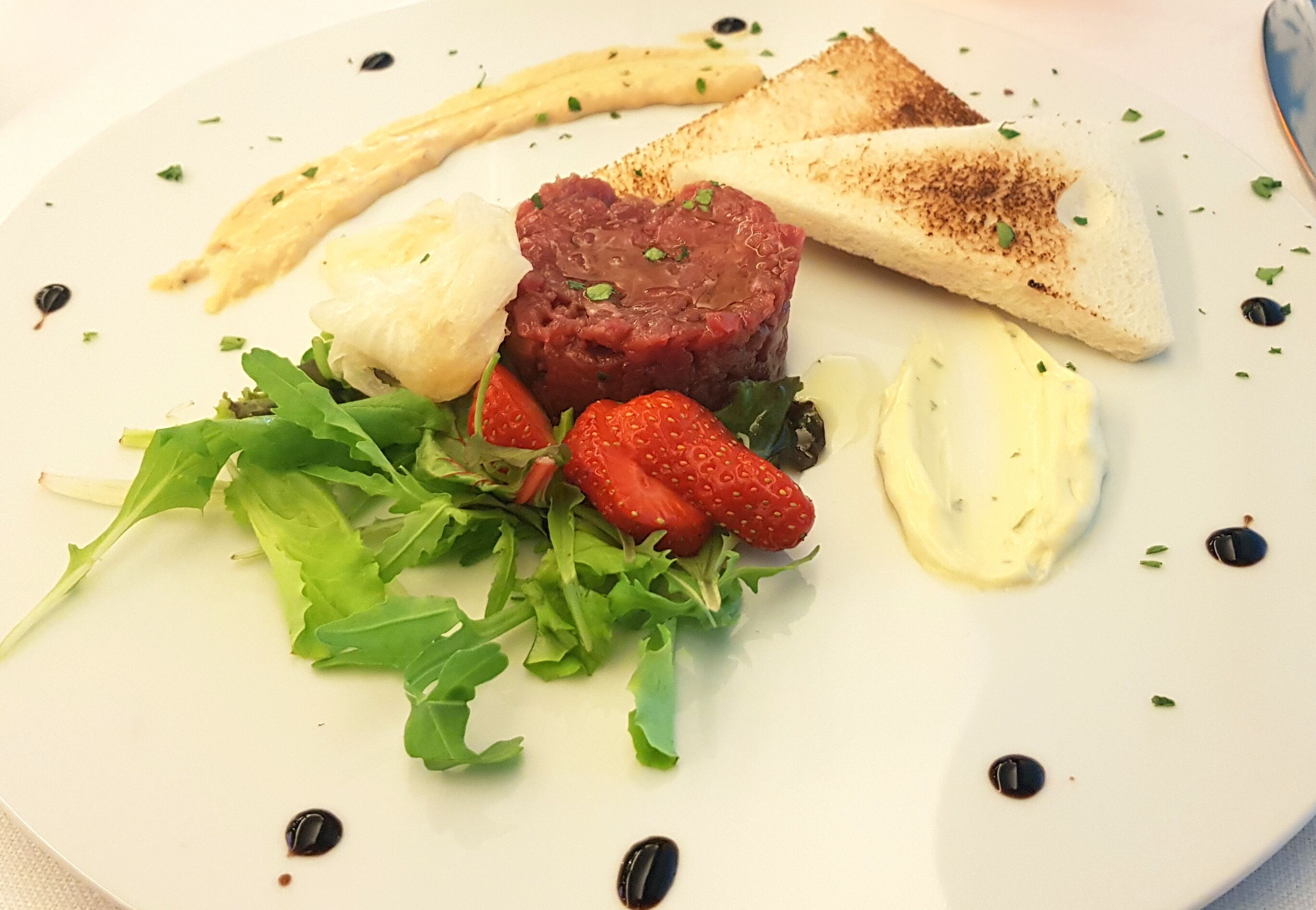 Une assiette comprenant du steak tartare, des toasts, des fraises et des légumes verts.