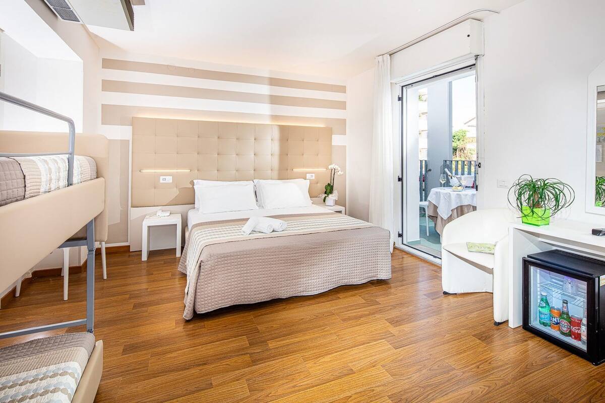 Una habitación de hotel moderna con una cama grande, un escritorio con computadora, un refrigerador pequeño y una puerta corrediza de vidrio al balcón.