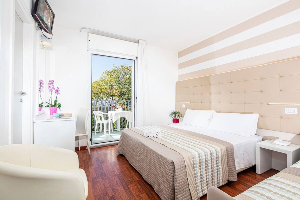 Una habitación de hotel moderna con una cama grande, una mesita de noche, una silla y un balcón con mesa y sillas.