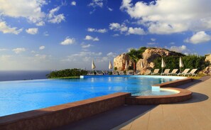 A pool with lounge chairs and umbrellas, a rocky cliff, and a view of the ocean.