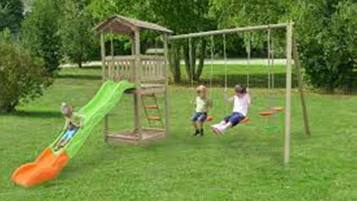 Children can play on the playground's slide and swings in a lush green space surrounded by trees.