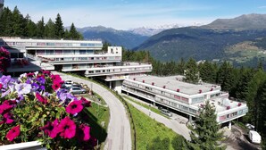 A modern hotel with multiple floors, outdoor seating, and a view of mountains. - Residence Sole Alto (Mezzana)