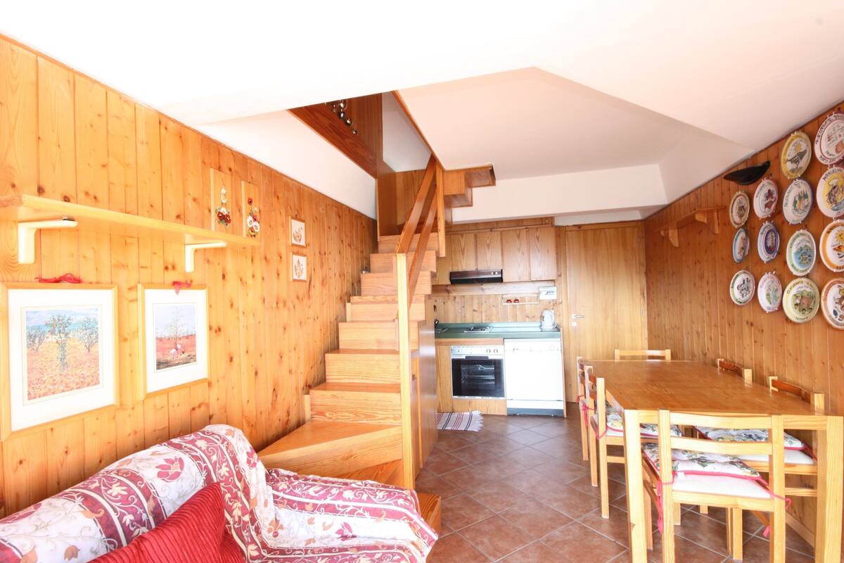 A wooden interior with a staircase leading to a kitchen, a dining area with chairs, and a sofa with patterned cushions.
