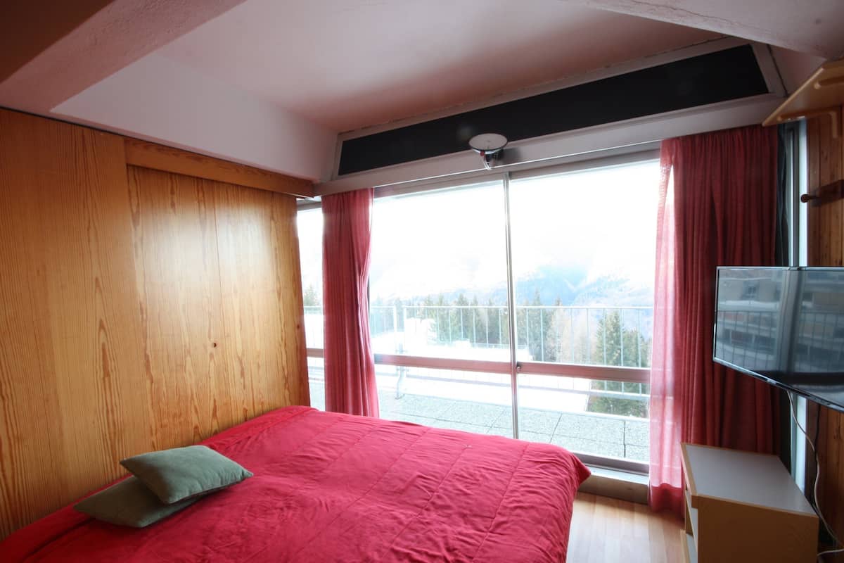 A bedroom with a large bed, wooden panel walls, a TV, and a balcony with a view of mountains.