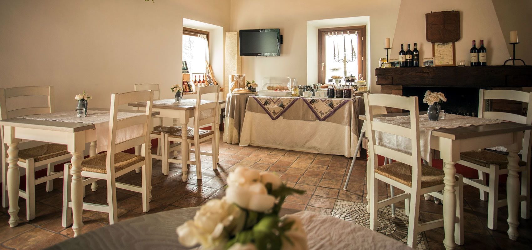 Un coin repas avec des chaises et des tables en bois peintes en blanc, un téléviseur fixé au mur et une cheminée ornée de bouteilles de vin sur le manteau.