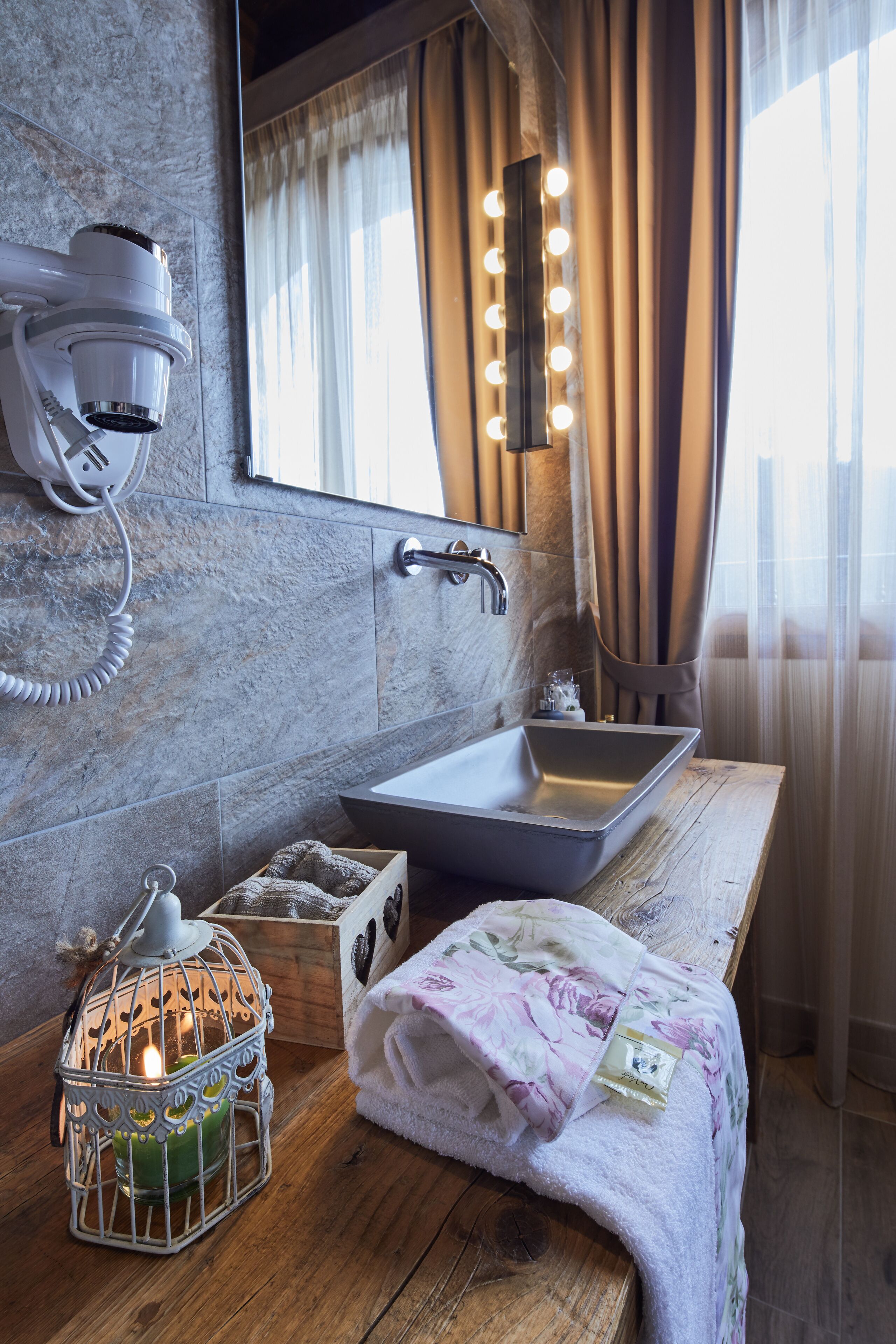 Un baño con pared de mármol, tocador de madera, lavamanos, espejo con luces, secador de pelo y una vela.