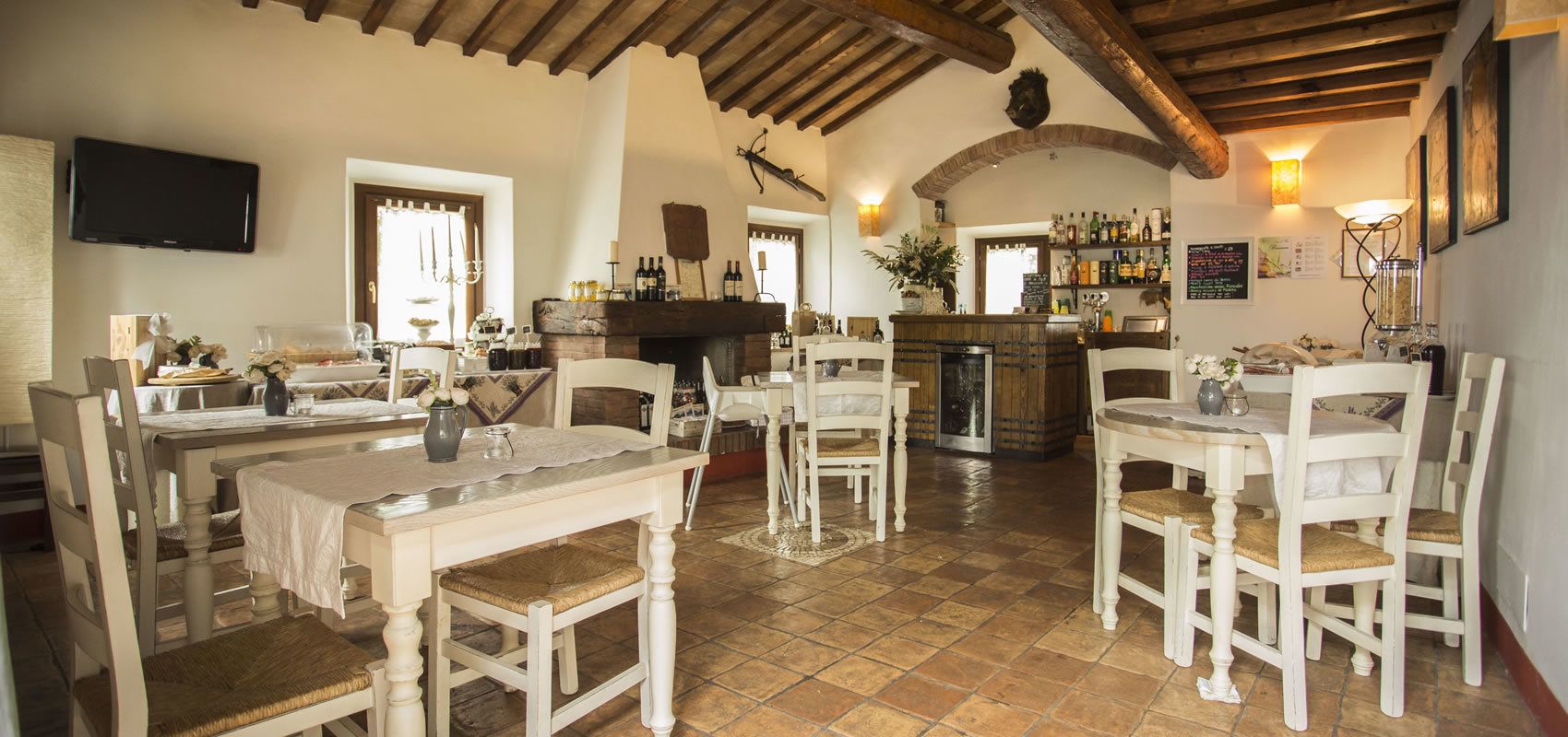 Un comedor con mesas y sillas de madera blanca, una chimenea y una zona de bar con botellas y vasos.