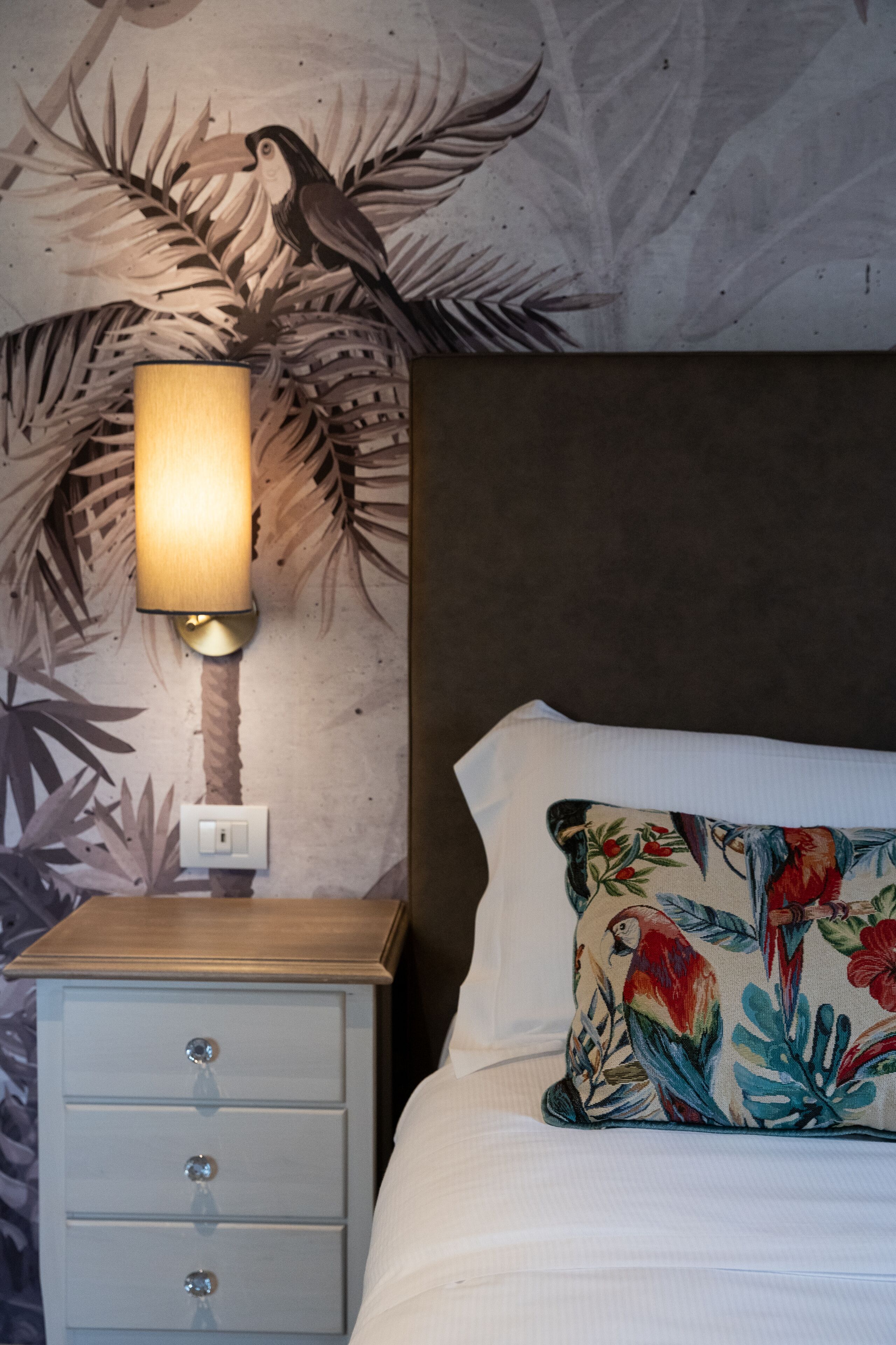 A bedroom with a nightstand, a lamp, a bed with a floral pillow, and a wall with a tropical mural.