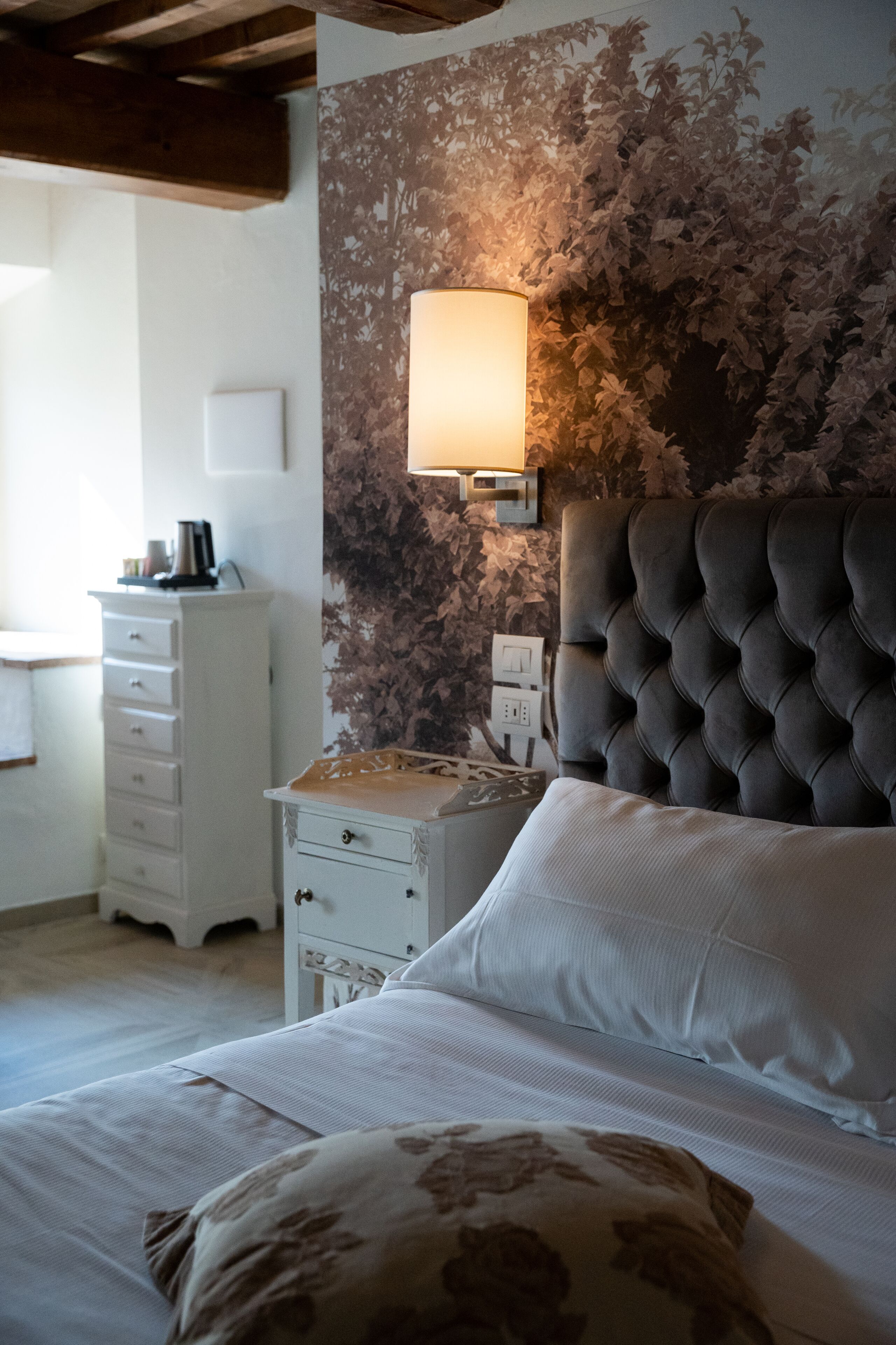 Une chambre à coucher avec une tête de lit capitonnée, une table de chevet avec une lampe, une commode et un coussin à motifs floraux.