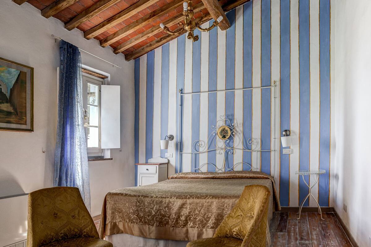 Une pièce avec un plafond en bois, un papier peint à rayures, un lit, deux fauteuils, une table de chevet et une lampe.