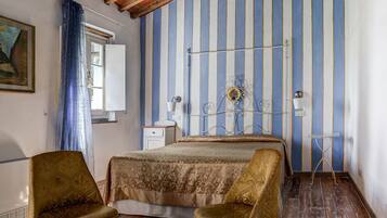 A room with a wooden ceiling, striped wallpaper, a bed, two armchairs, a bedside table, and a lamp.