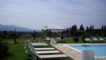 Zona de piscina con sillones de descanso y una pérgola, rodeada de vegetación y montañas al fondo.