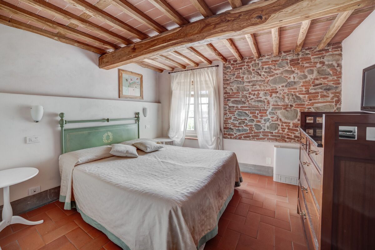 Una camera da letto con parete in pietra, soffitto in legno, un letto con testiera verde, un comodino bianco e un piccolo termosifone bianco.