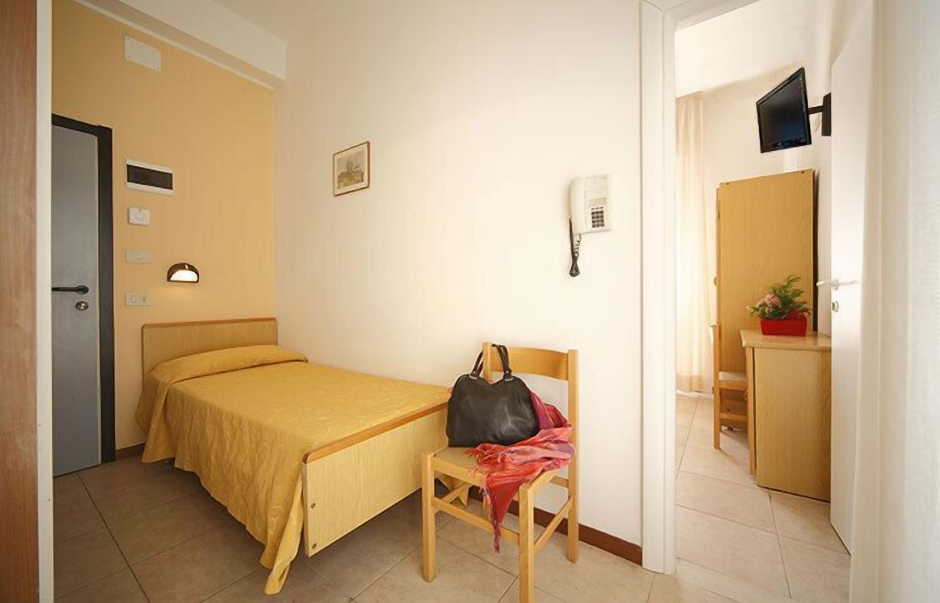 A small, neatly arranged bedroom with a single bed, a chair, a desk, a telephone, and a television.