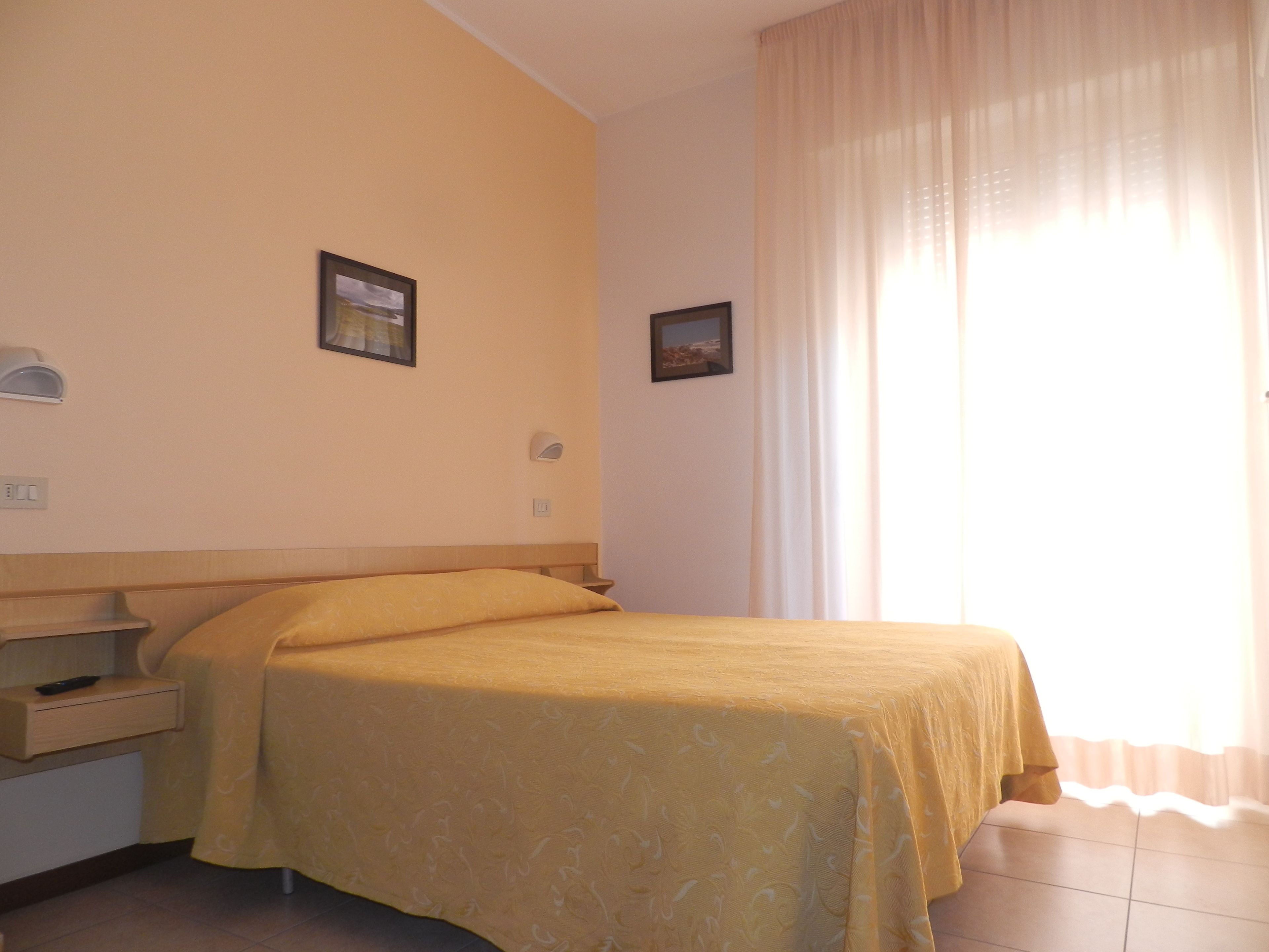 A hotel room with a bed, a nightstand, a wall-mounted lamp, and a window with curtains.
