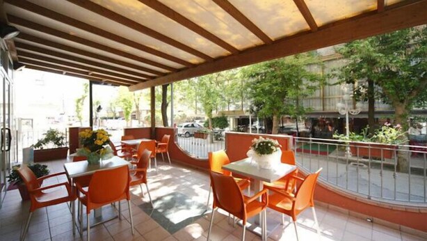 An outdoor cafe with orange chairs and tables, a covered area with wooden beams, and a view of the street and greenery. - Hotel Sport (Rimini)