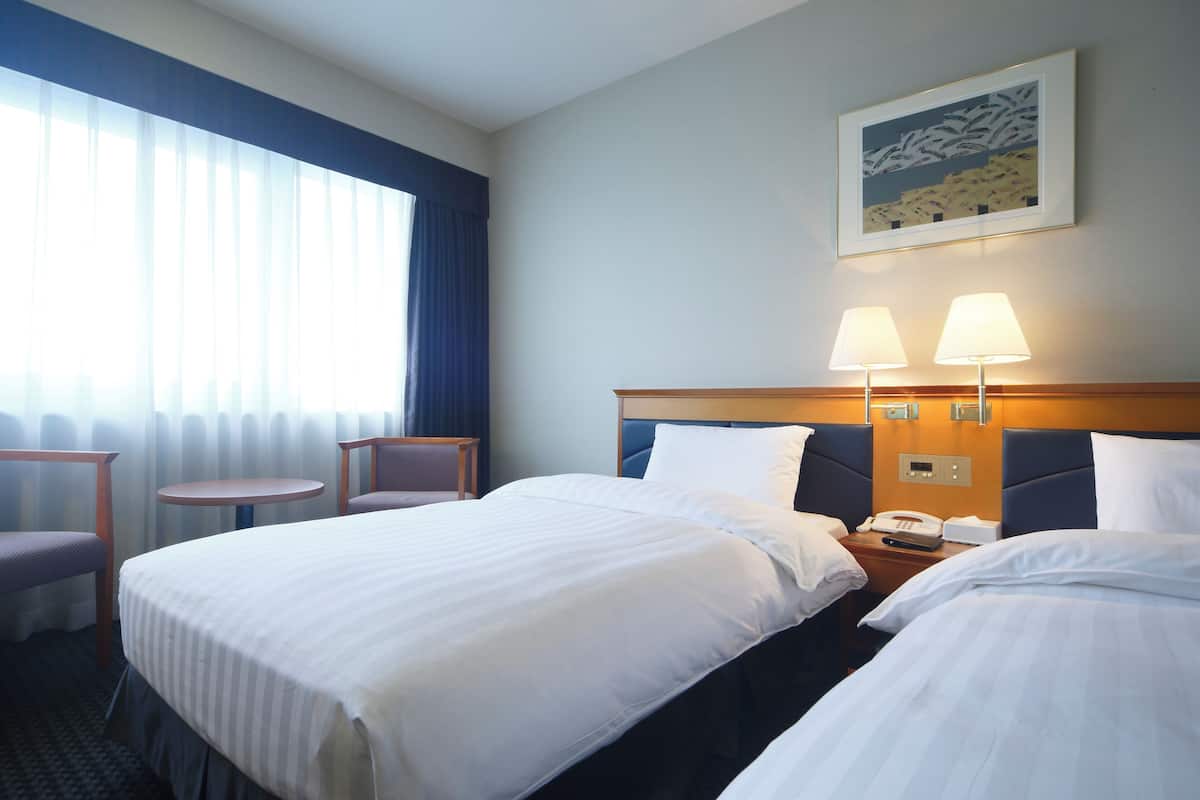 Habitación de hotel con dos camas, una mesita pequeña, dos sillas, una mesita de noche con un teléfono y un cuadro en la pared.