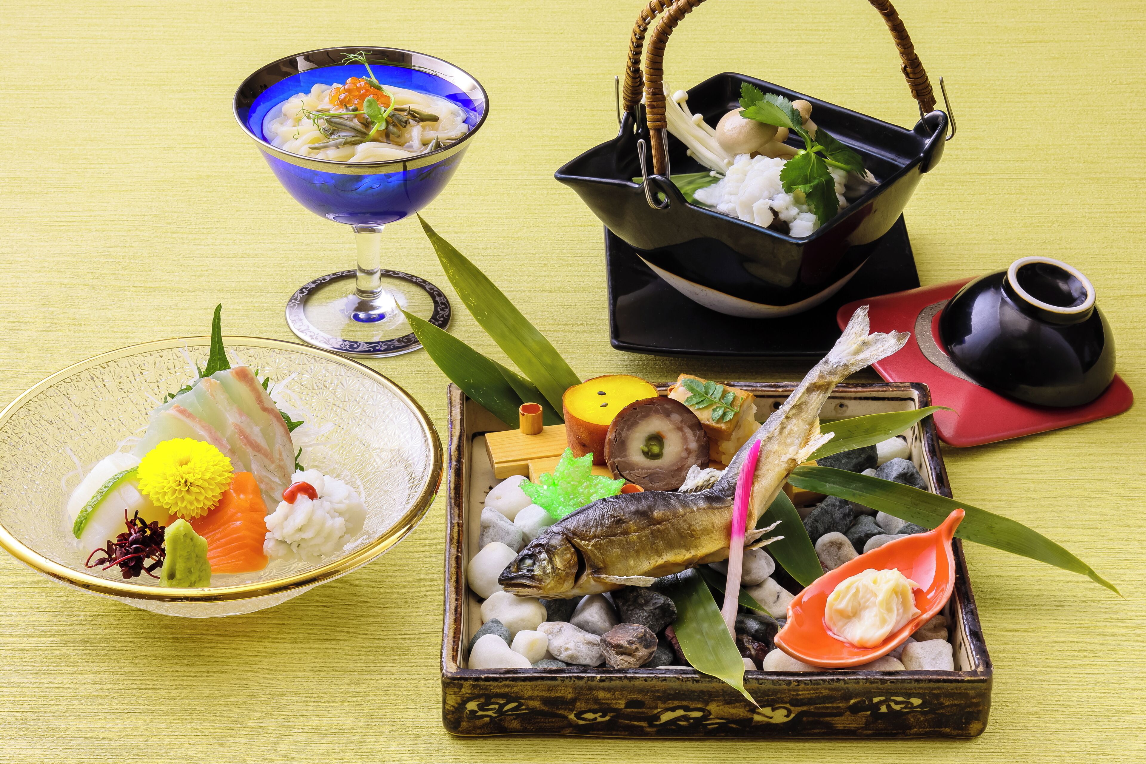 Variedad de platos japoneses, como sushi, sashimi y una escultura de pescado, presentados sobre una mesa con fondo amarillo.