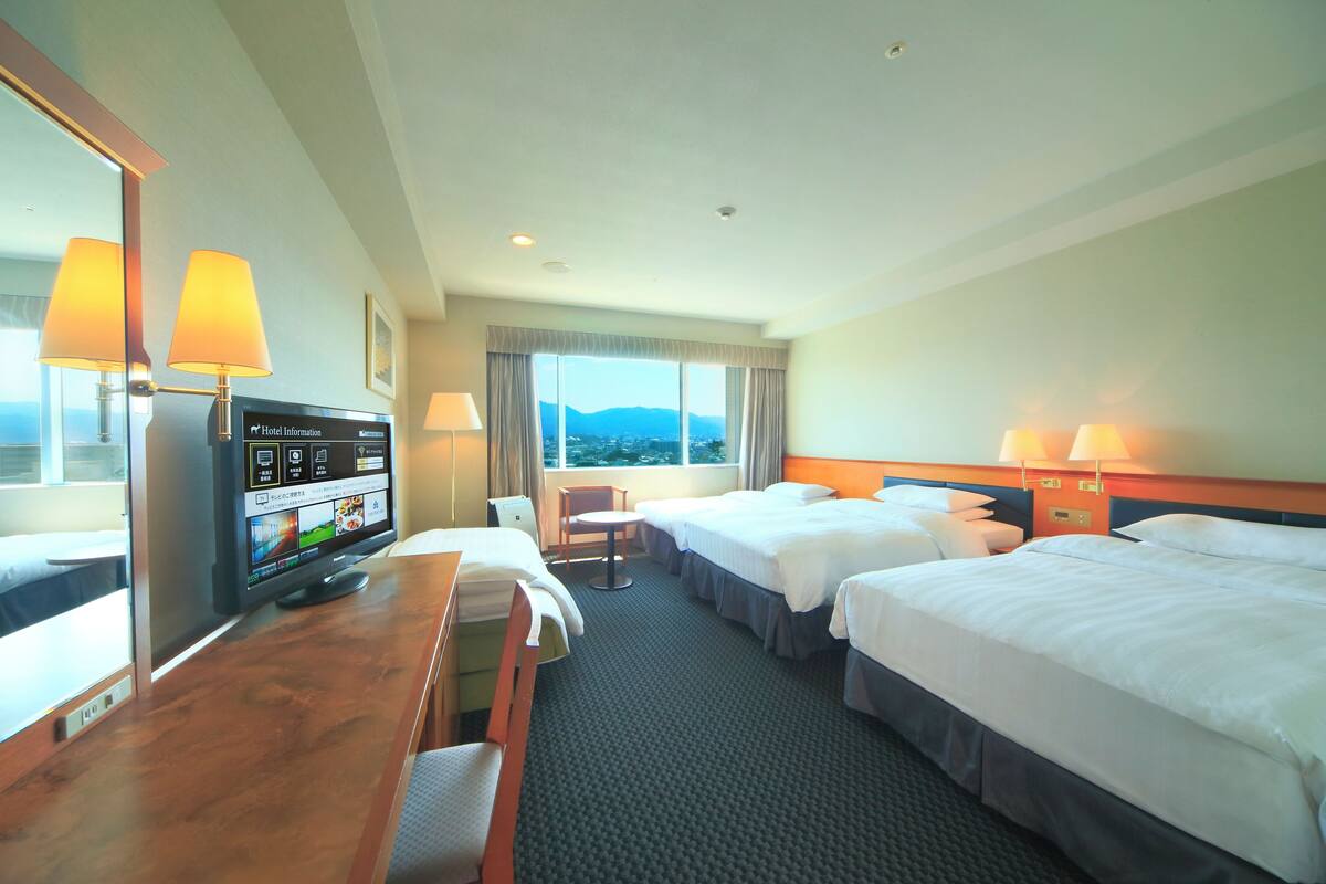 Habitación de hotel con dos camas, un escritorio, televisión y ventana con vista a las montañas.