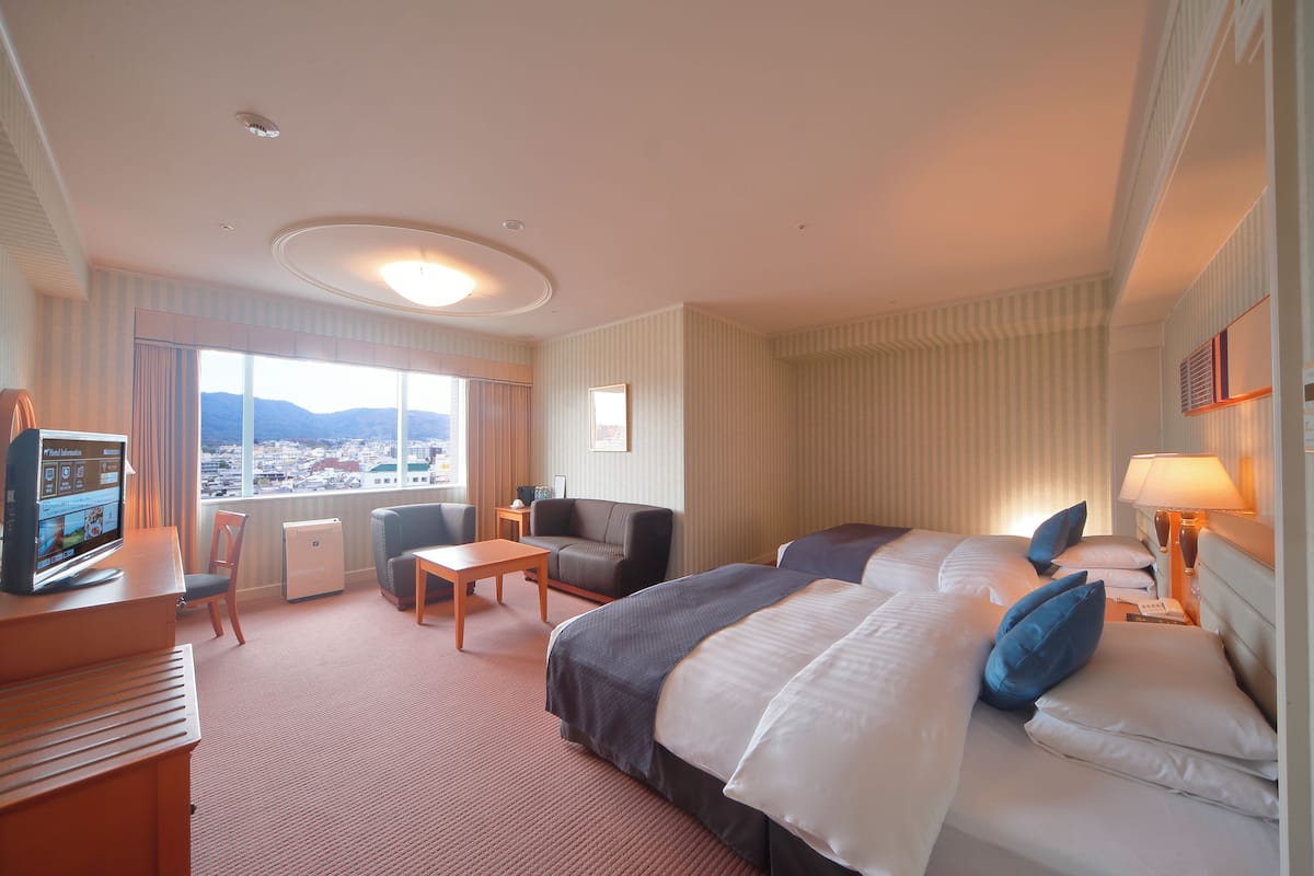 Habitación de hotel con una cama grande, un escritorio con computadora, una silla, una mesita y vistas a un paisaje urbano a través de amplios ventanales.
