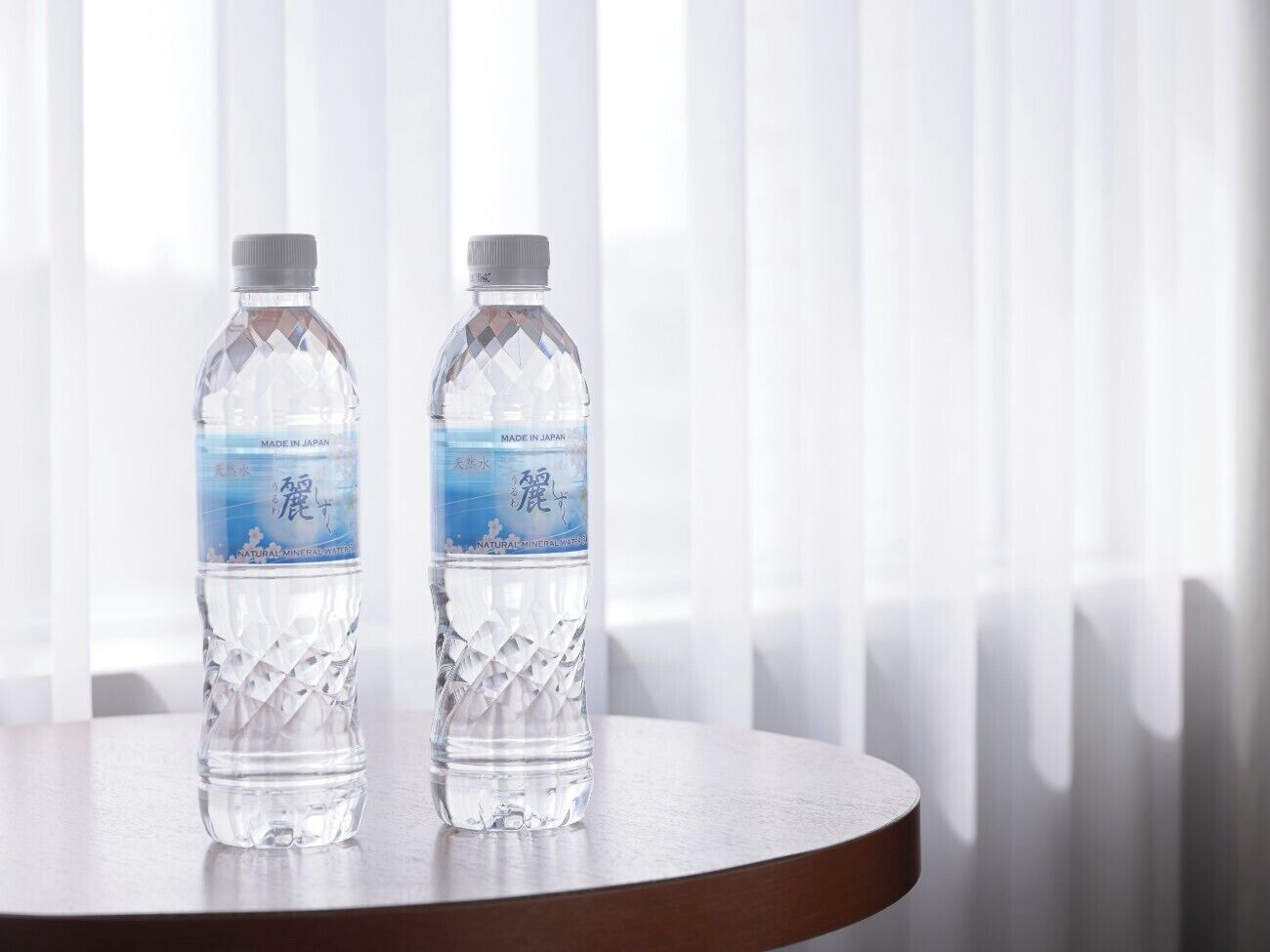 Two water bottles on a round table with a wooden base, placed in front of a window with vertical blinds.