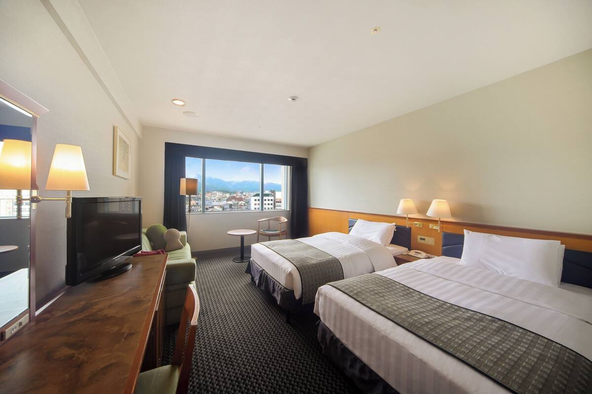 Habitación de hotel con dos camas, un escritorio con televisor, una silla, una mesita y una ventana con vista a la ciudad.