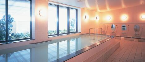 A modern indoor swimming pool with clear glass walls, multiple shower stations, and a tiled floor.