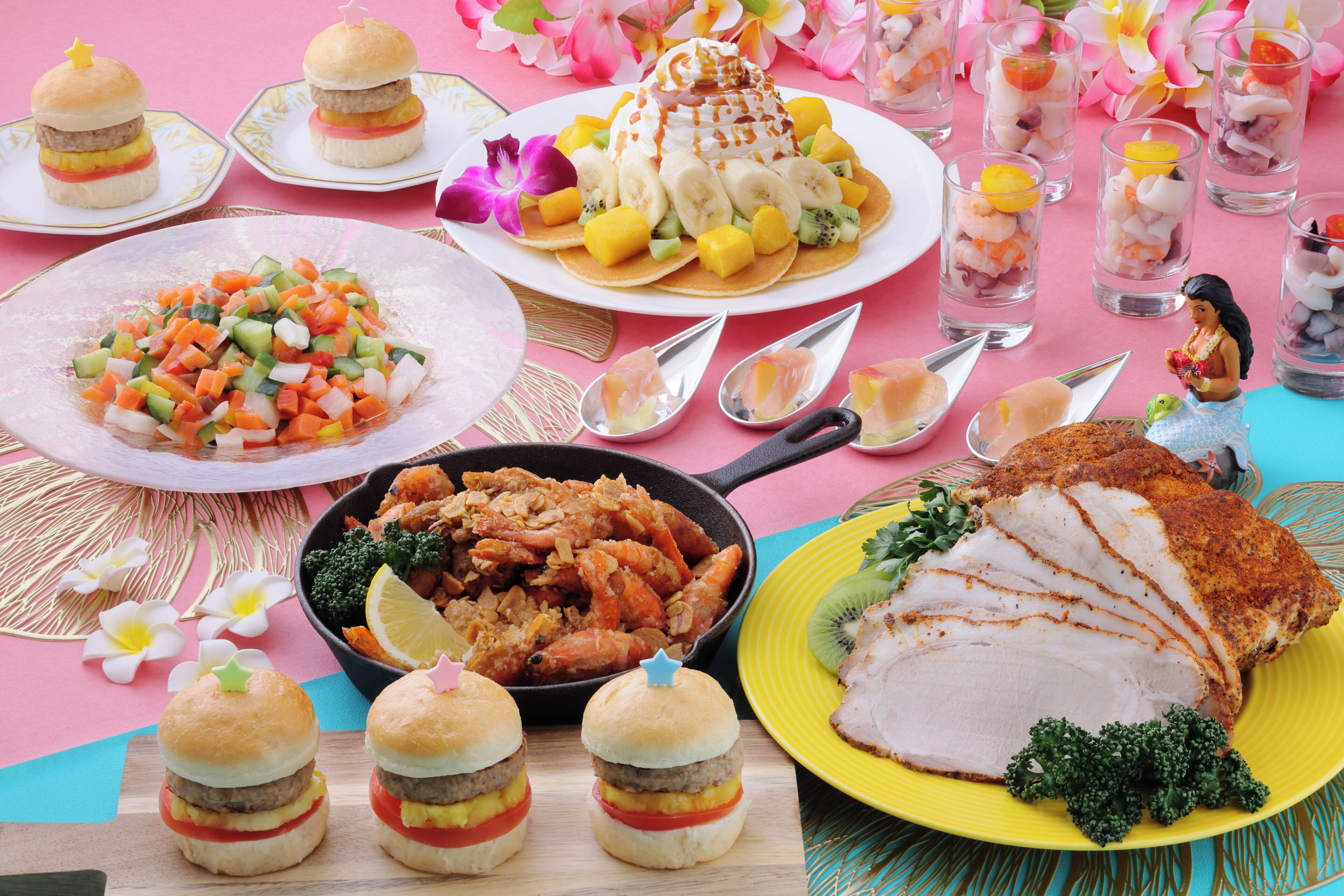 A variety of dishes including burgers, a fruit dessert, and a roasted chicken dish, arranged on a table with a pink and blue tablecloth.