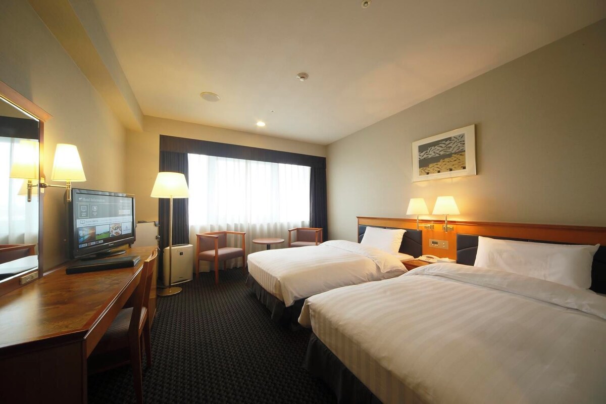 A hotel room with two beds, a desk with a computer, a chair, and a window with curtains.