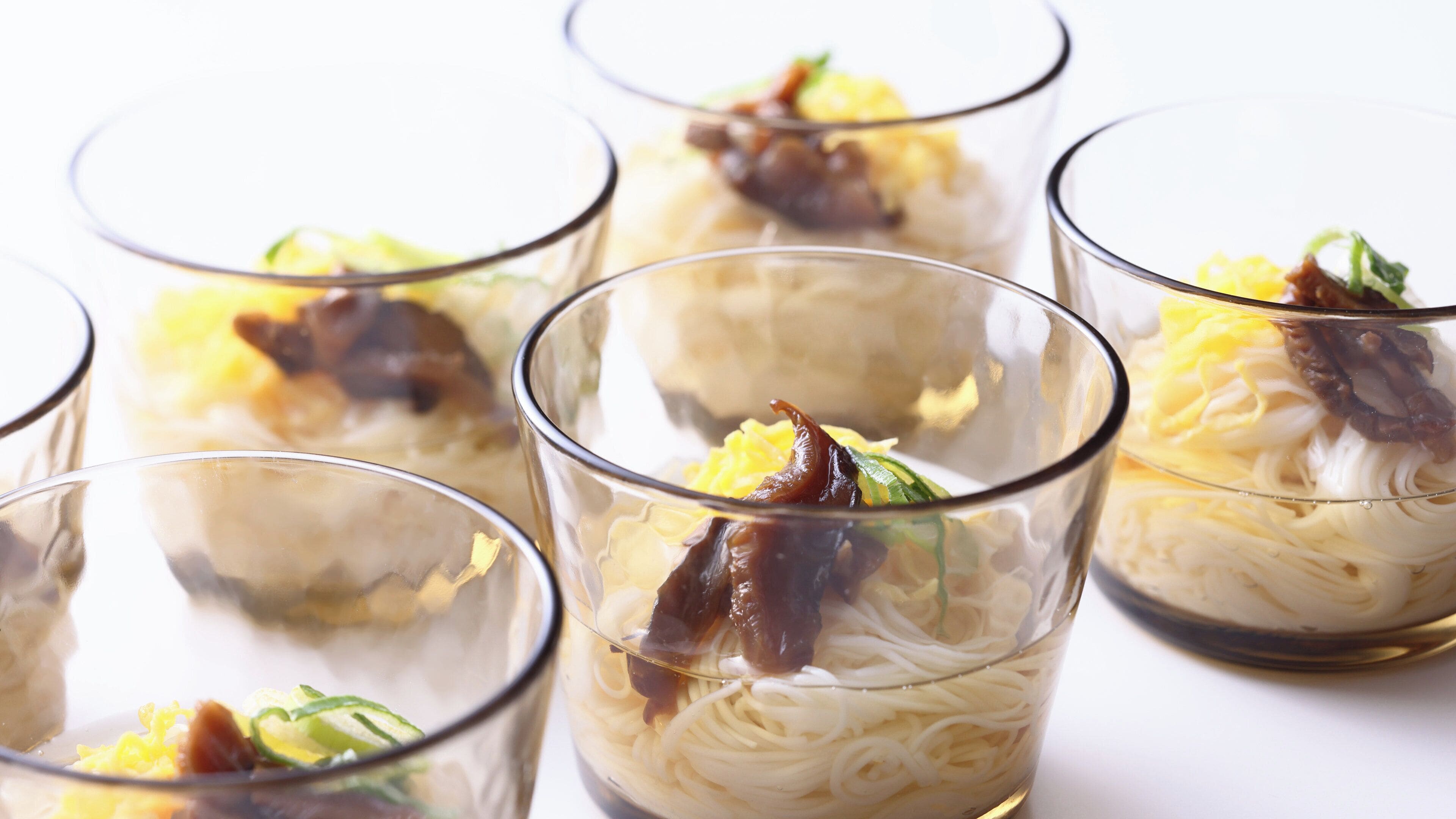 A series of clear glass cups containing a noodle dish with vegetables and meat.