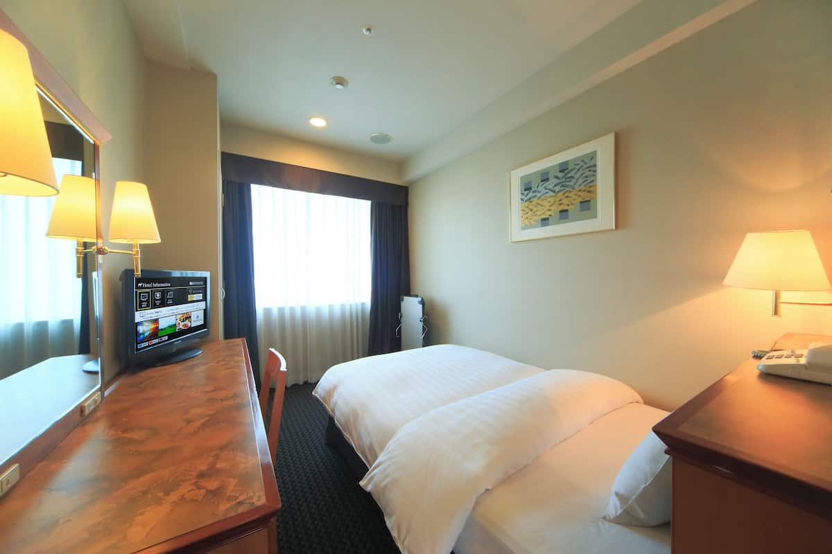 Una habitación de hotel con cama, escritorio, televisor mostrando información del hotel y ventana con cortinas.