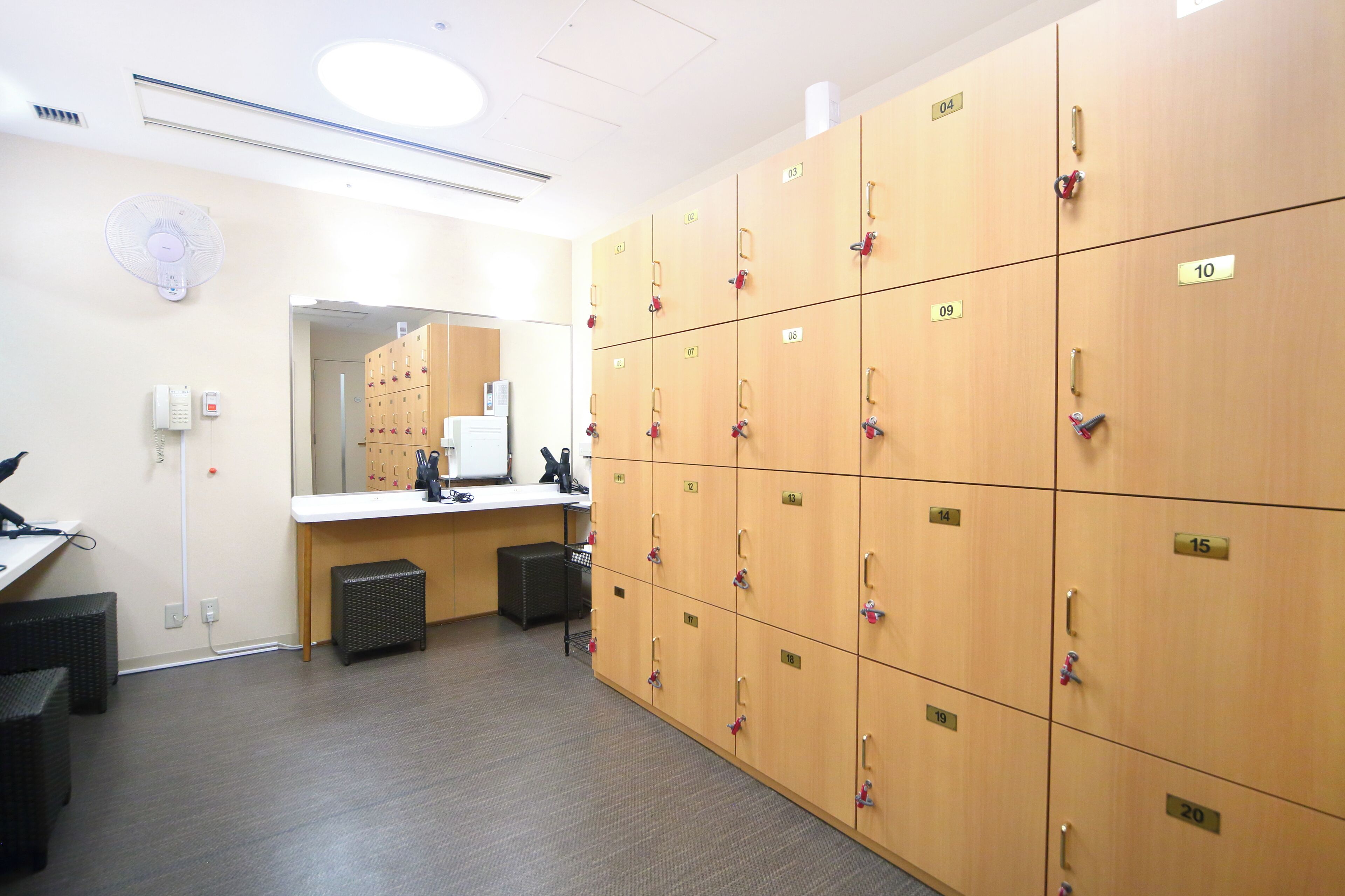 A locker room with multiple wooden lockers, a mirror, a desk with a computer, and a fan.