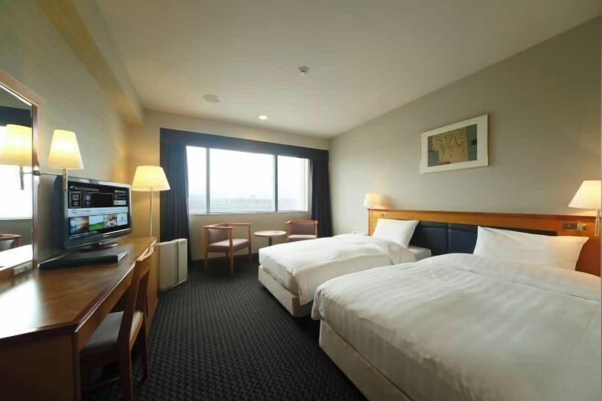 Habitación de hotel con dos camas, un escritorio con televisor, una silla, una mesita y ventana con vistas.