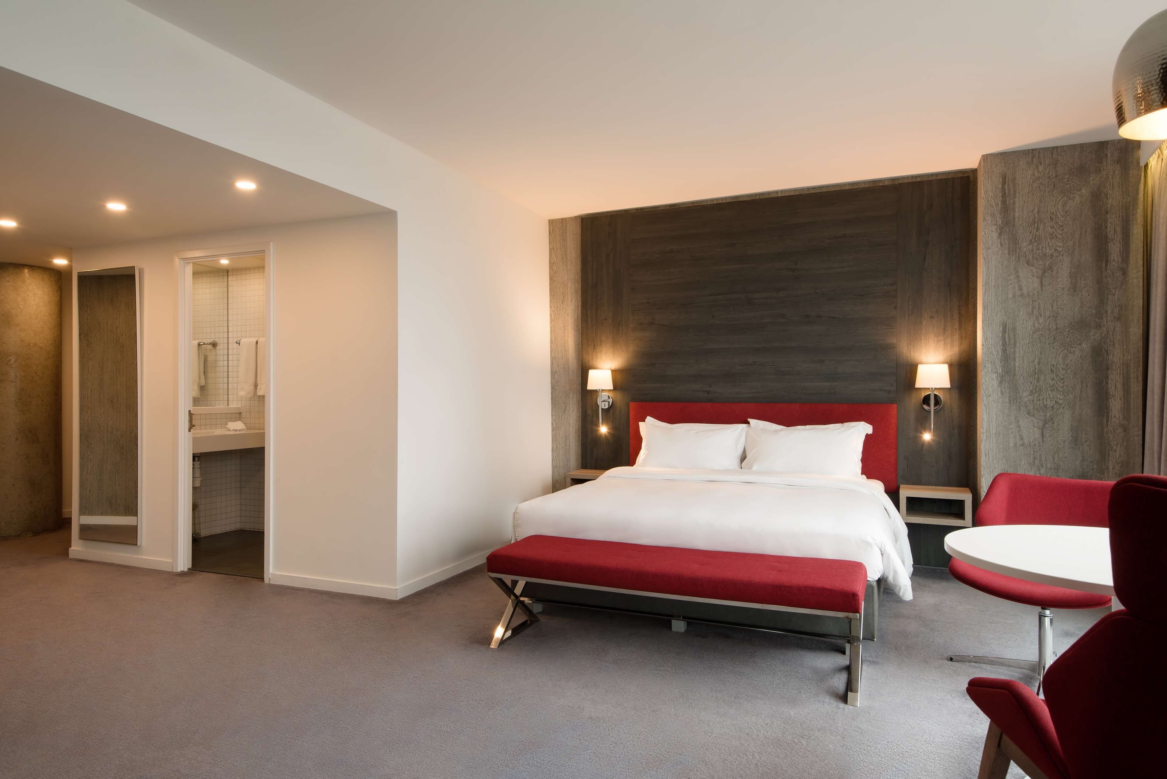 Una habitación de hotel moderna con una cama grande, un banco rojo, una mesita pequeña y un área de ducha.