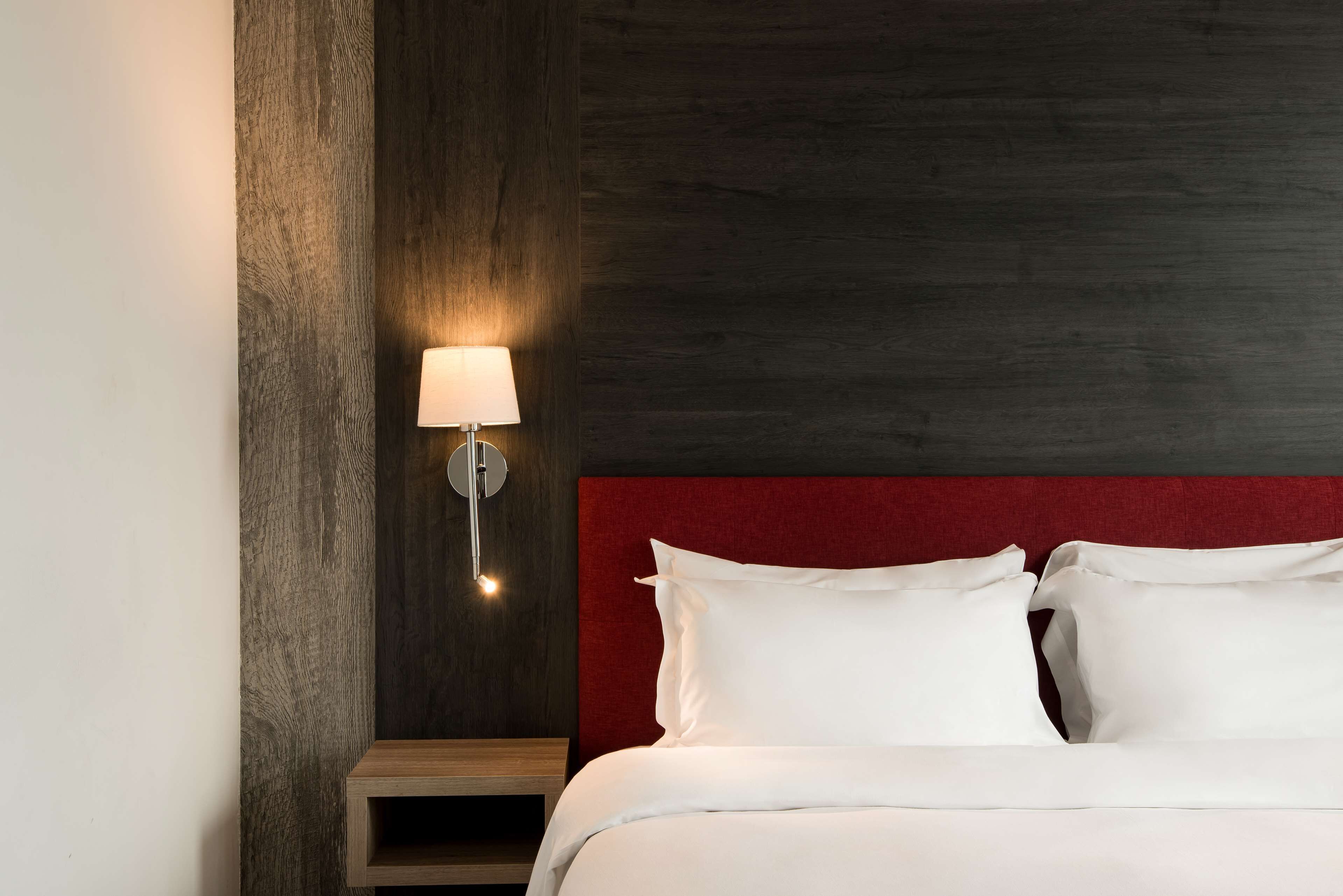A hotel room with a wooden accent wall, a red headboard, and a bedside lamp.
