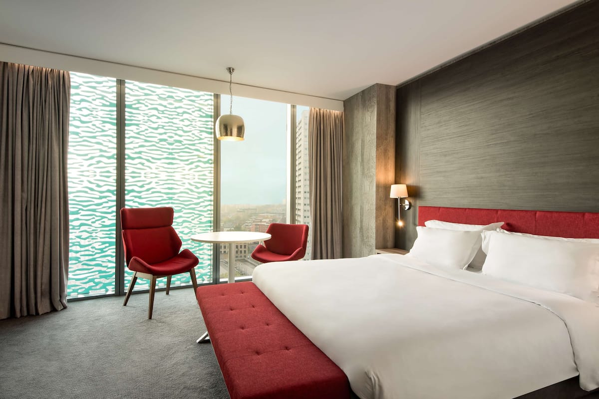A hotel room with a large bed, a red chair, a small table, and a view of the city through large windows.