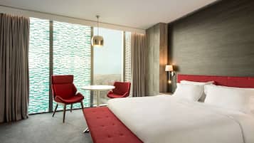 Quarto de hotel com uma cama grande, uma cadeira vermelha, uma mesa pequena e vista da cidade através de amplas janelas.
