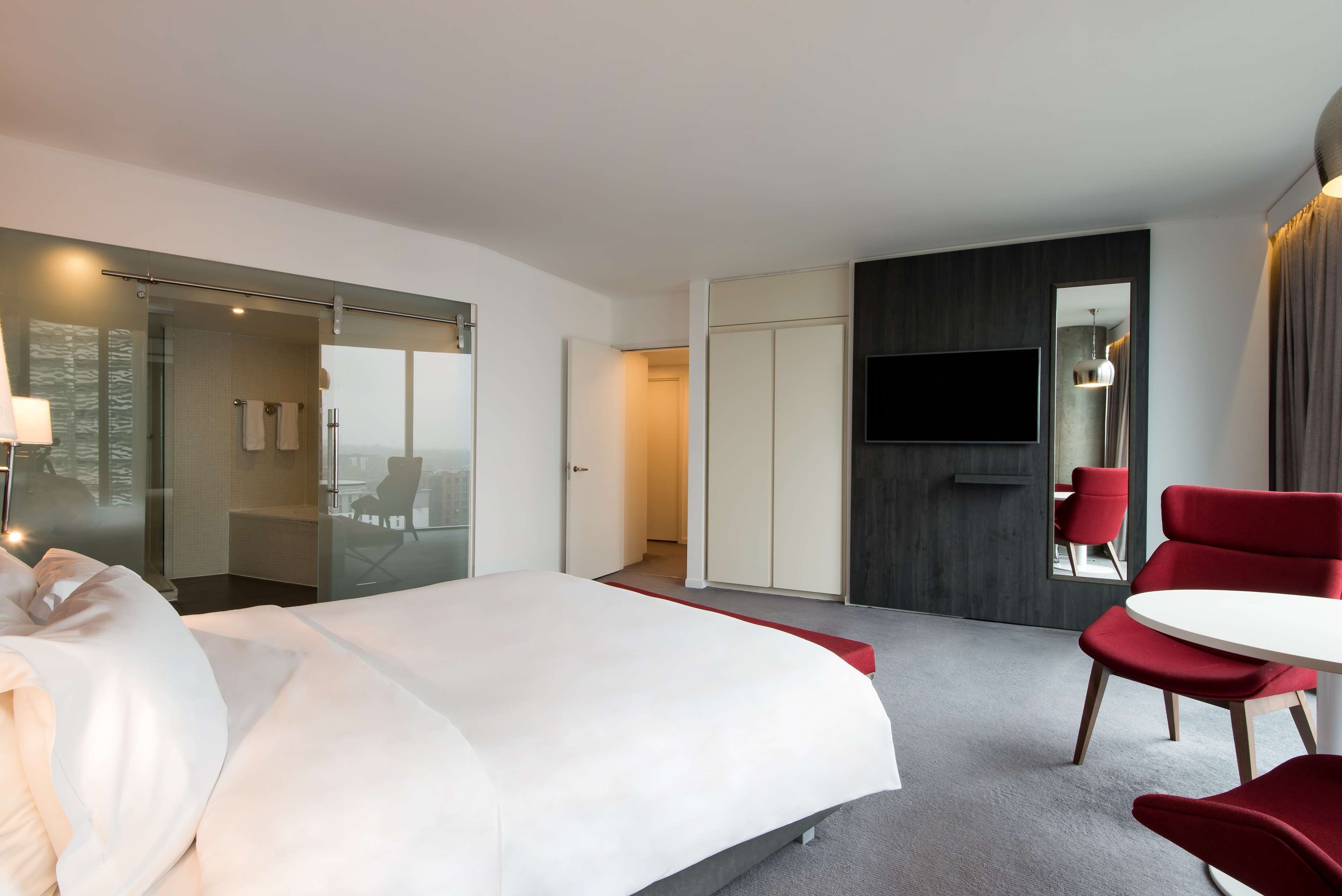 Une chambre d’hôtel avec un lit, une télévision, un fauteuil rouge et une porte en verre donnant sur une salle de bain.