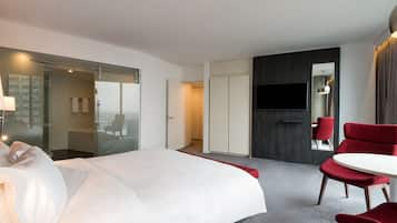 A hotel room with a bed, a TV, a red chair, and a glass door leading to a bathroom.