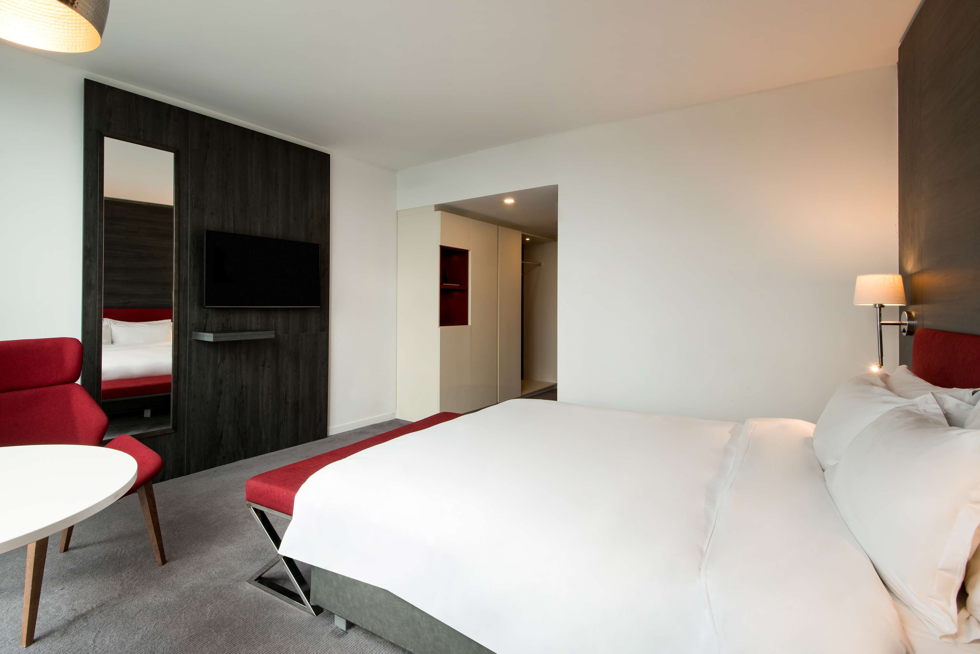 A modern hotel room with a large bed, a TV mounted on the wall, a red chair, and a small table.