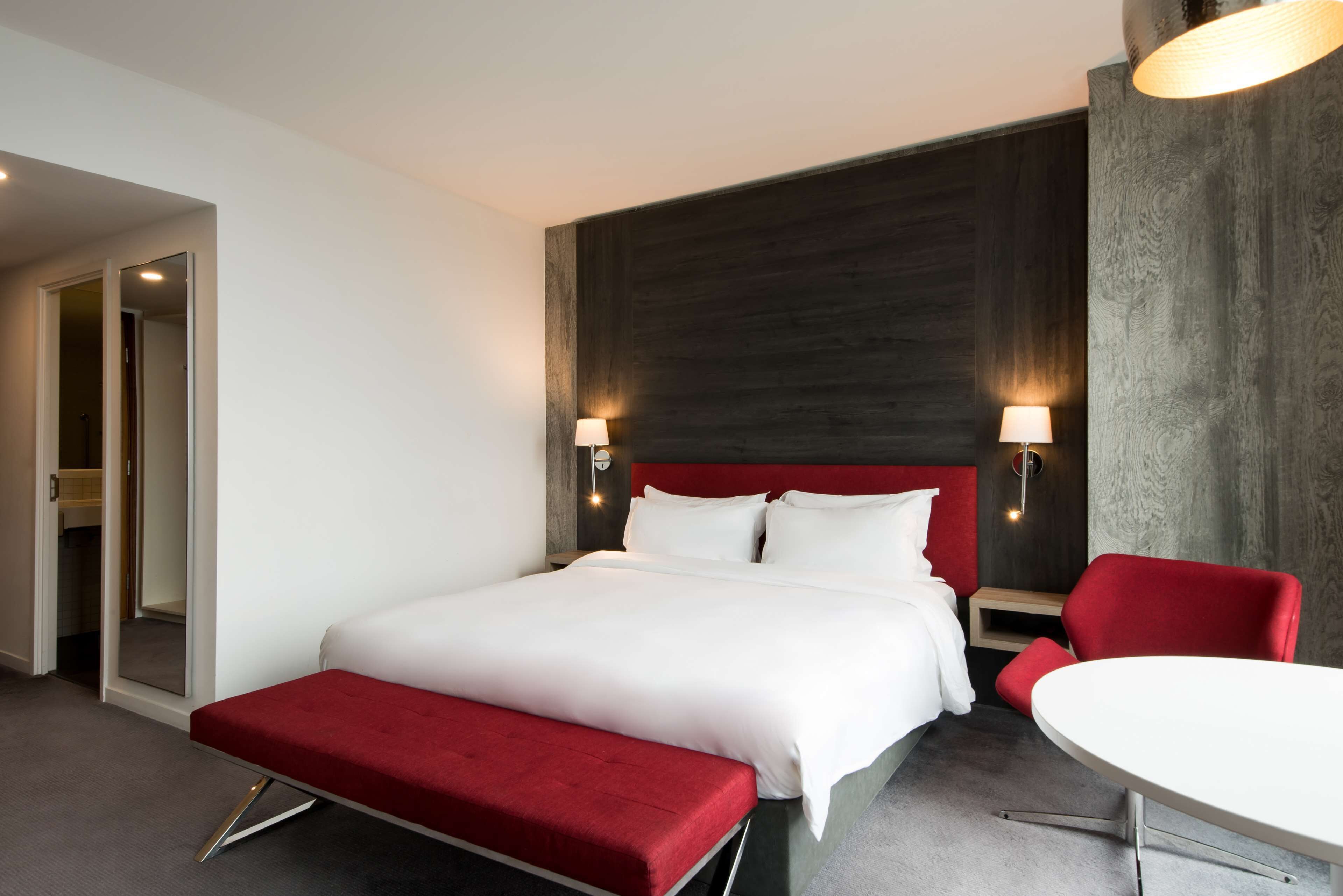 A hotel room with a large bed, a red bench, a white table, and two wall-mounted lamps.