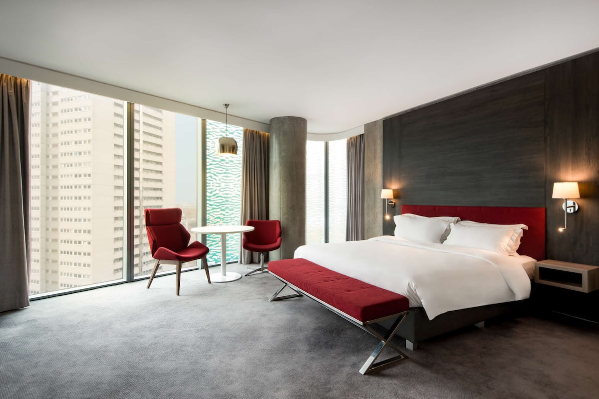 A modern hotel room with a large bed, a red bench, two red chairs, a small table, and a large window offering a city view.