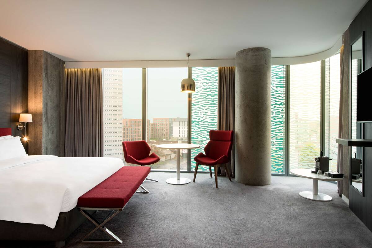 A hotel room with a bed, a red bench, two red chairs, a small round table, a large window with a view of buildings, and a large pillar.