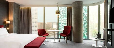 A hotel room with a bed, a red bench, two red chairs, a small round table, a large window with a view of buildings, and a large pillar.
